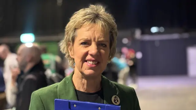 A woman with short hair holding a blue clipboard. She is wearing a green blazer and smiling at the camera.
