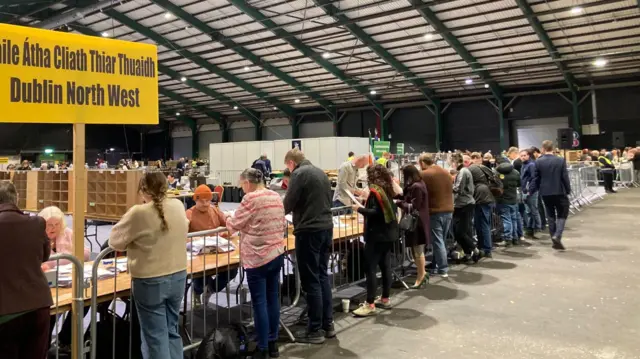 A crowd of people stand at metal barriers, watching other people sort through pieces of paper at tables. There is a large yellow sign which says 'Átha Cliath Thiar Thuaidh - Dublin North West'.