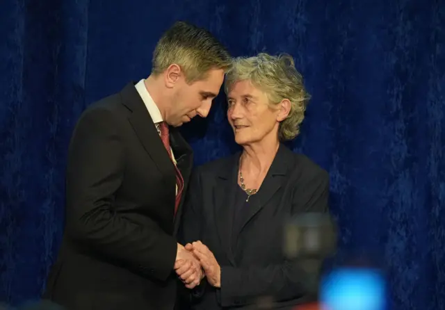 Catherine Connolly and Simon Harris are shaking hands. There's a dark blue backdrop behind them.