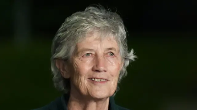 Catherine has short grey hair and is smiling at the camera as she wears a dark green coat. The background is blurred.
