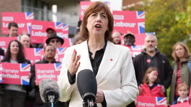 Lucy Powell in white blazer and white top stands in front of a group of supporters holding signs reading Renew Britain
