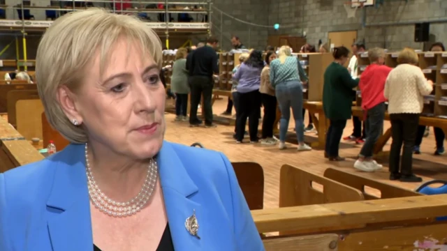 Humphreys is wearing a blue jacket with a leaf pin and a pearl necklace. People are standing counting votes in the background in a hall.