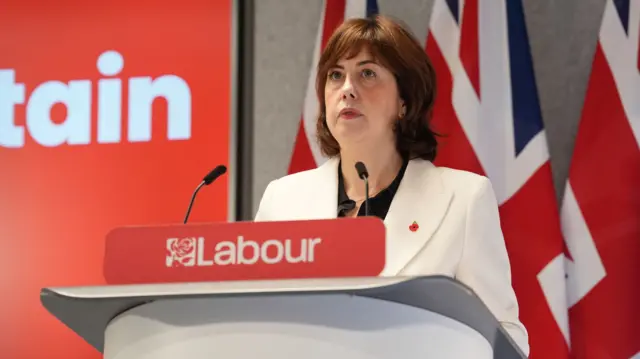 Lucy Powell in white blazer and black top stands behind a grey podium, three Union flags behind her