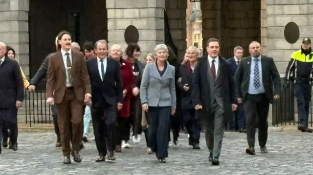 Catherine Connolly walking in the courtyard of Dublin Castle, surrounded by a group of men and women