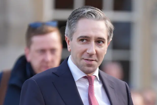 Harris is wearing a navy suit with a white shirt and red tie. The background is blurred. He's outside.
