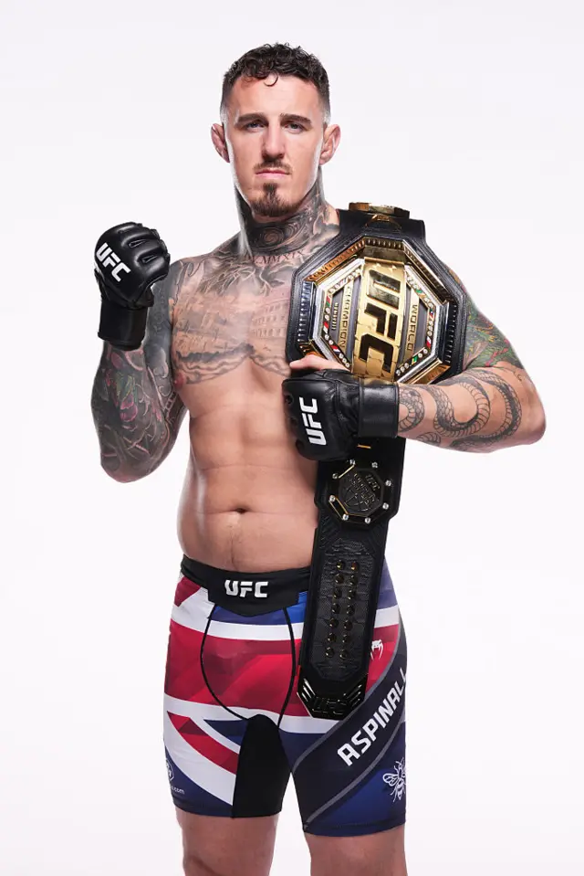 Tom Aspinall wearing Union Jack UFC shorts and holding the UFC heavyweight title over his shoulder