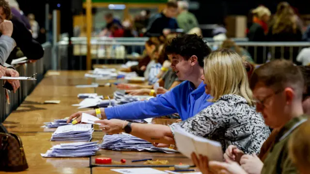 People siting at a table counting votes. There is a silver barrier around the table with people hanging over with clipboards.
