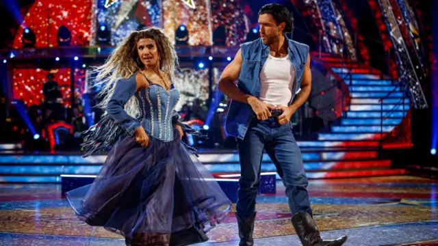 Balvinder wears her ombre hair in curly half pigtails, and has on a denim rhinestone corset and denim shawl with tassels