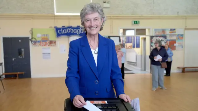 Catherine Connolly, with short grey hair, wearing a white top and a blue blazer. She is standing in a school putting her vote into a ballot box.