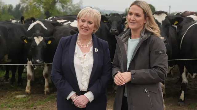 Heather Humphreys (left) with Helen McEntee on a farm in county Meath - they are standing in front of a field with black and white cows in it.