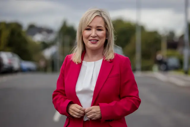 O'Neill is standing outside. She's wearing a pink suit with a white top.