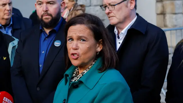 Mary Lou McDonald has short brown hair and is wearing a green coat and leopard print top as she speaks to the media.