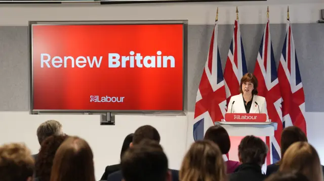 Wide shot of Lucy Powell delivering her speech from a low stage. She's in a white blazer and black top standing behind a grey podium, a group of people sitting in front of her. Behind her are four Union flags, and a wall-mounted TV screening the wording Renew Britain on red background