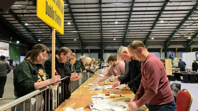 A long table with people lined up on either side, counting ballot papers that are scattered on the table.