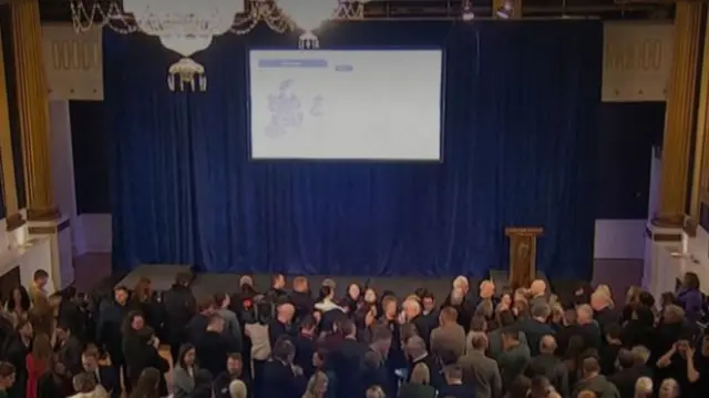 A hall with a lot of people standing in it awaiting declaration. There is a large screen showing results.