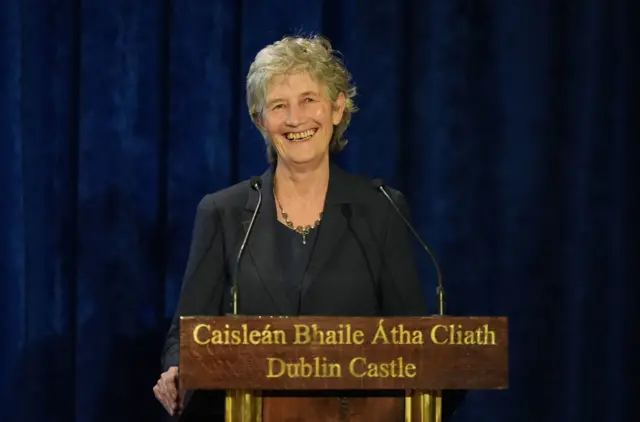 Connolly is standing behind a mic. She's smiling. She's wearing a dark suit and there's a dark blue backdrop behind her.