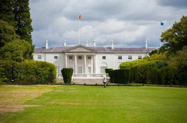 The Áras an Uachtaráin. A cyclist in on the lawn leading up to the building. It's cloudy.