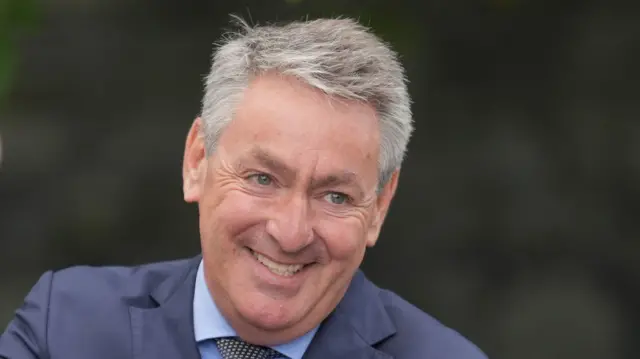 Kelleher has short grey hair and is smiling in the picture with a blue suit, shirt and patterned tie.