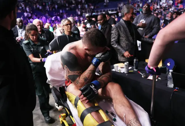 Tom Aspinall holds his hand to his face as he sits on a stretcher