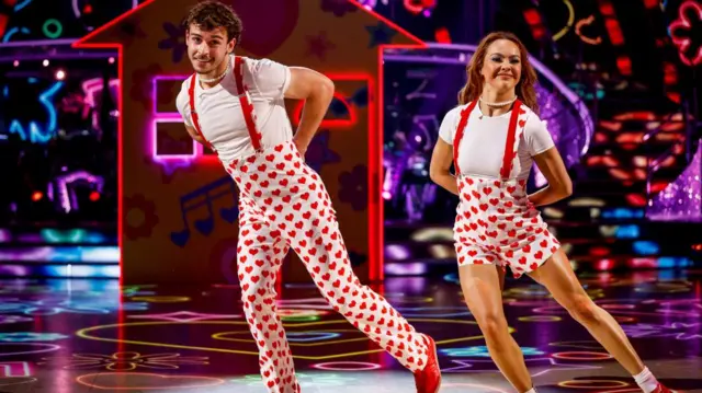 George Clarke wears white high-waisted heart print white trousers with suspenders, adidas trainers and a pearl necklace