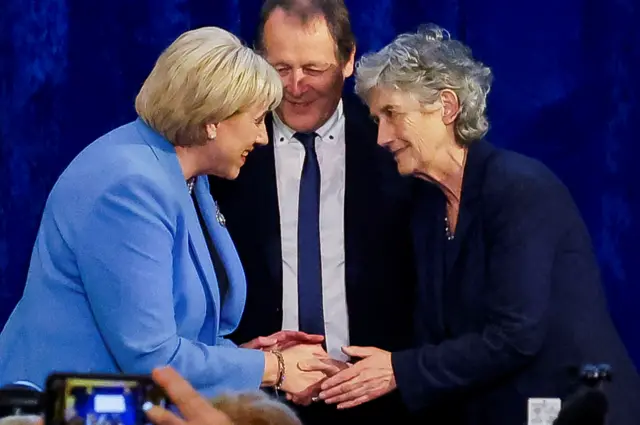 Heather Humphreys and Catherine Connolly shaking hands, next to Connolly's husband. Humphreys has shoulder length blonde hair and is wearing a blue blazer. Connolly has short grey hair and is wearing a navy blazer, while her husband wears a black jacket, white shirt and blue tie.