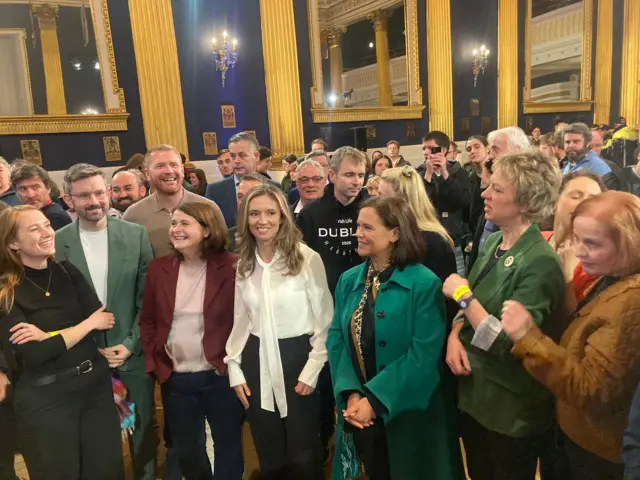 Holly Cairns, Iva Bacik and Mary Lou McDonald are standing together. There are crowds around them. They're in a hall.
