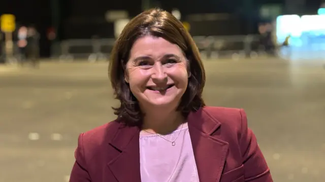 A woman with shoulder-length brown hair stands smiling at the camera. She is wearing a pink top and a maroon blazer.