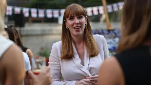 Rayner in a creme shirt, smiling as she holds a glass