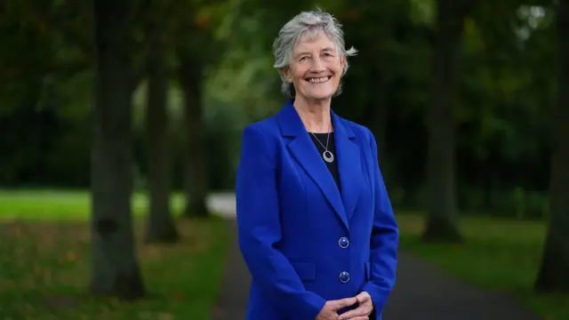 Catherine Connolly, standing in a park, smiling at the camera. She has short grey hair and is wearing a black top and blue jacket.