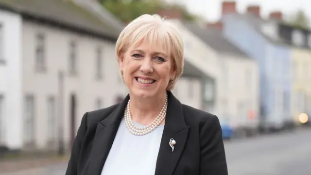 Heather Humphreys has short blonde hair and is smiling at the camera as she wears a black blazer, white top and pearl necklace.