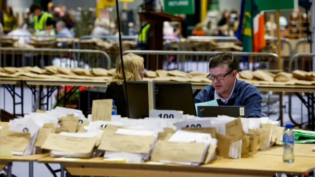A man sitting at a table with piles of ballot papers on it