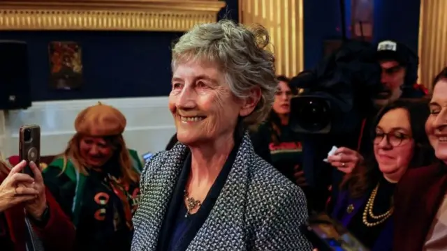 Catherine Connolly has short grey hair and is smiling as she walks through a hall at Dublin Castle. She is wearing a black and white coat.