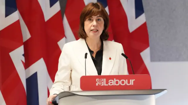 Lucy Powell stands behind podium, four Union flags behind her