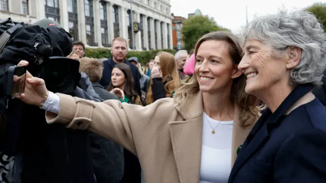 Holly Cairns, a woman with long brown hair, wearing a white told and beige jacket, standing beside Catherine Connolly, a woman with short grey hair, wearing a navy jacket. Cairns is holding out her phone and the pair are taking a selfie. There is a group of people behind them.