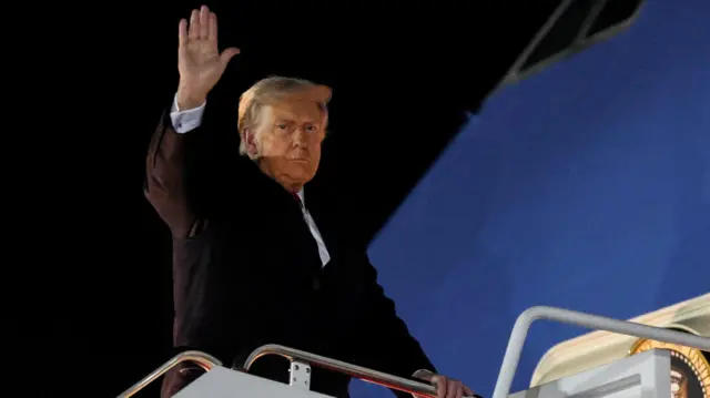 US President Donald Trump waves as he boards Air Force One, wearing a dark suit jacket.