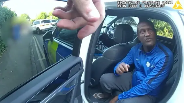 Police body-cam footage of Hadush Kebatu sitting in a police patrol car wearing a blue Nike top, track trousers and white flip flops