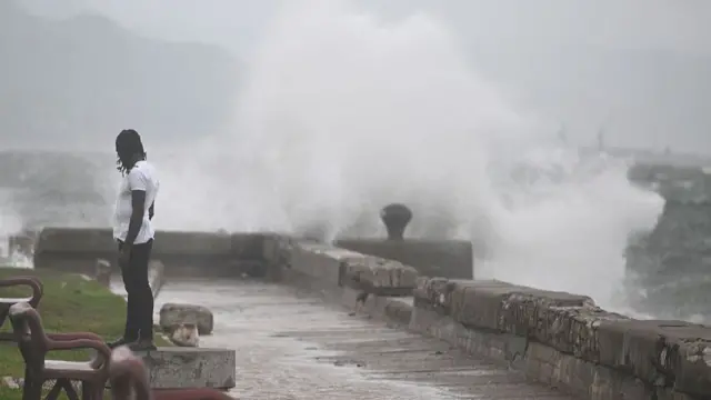 A man stands on a wall of waterfront watching big waves crash into the bricks