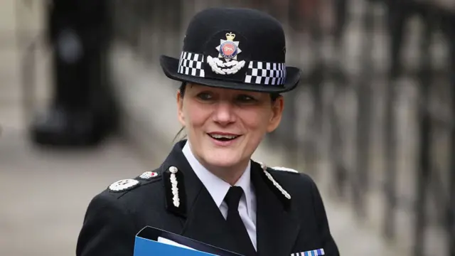 Dame Lynne Owens is looking to the side of the camera, wearing her police uniform and smiling. She is carrying a blue folder.
