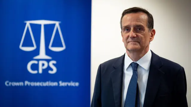 Stephen Parkinson to the right of the frame wearing a dark suit and blue tie. Behind him is a panel with the symbol of the Crown Prosecution Service (a simplified white scale on blue background, CPS written below the base)