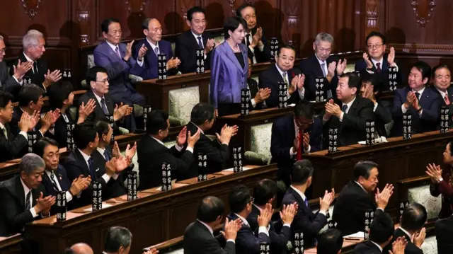 Takaichi standing up in a purple blazer. Around her, seated in rows are men in black suits clapping