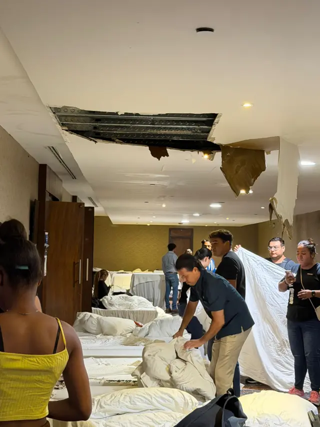 A group of people stand around handling bed linen in the room of a hotel. A hole gapes in the roof, and plasterboard hangs down.