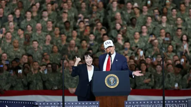 Takaichi giving a thumbs up as she stands beside Trump at a podium. Behind them is a sea of people in green military uniform