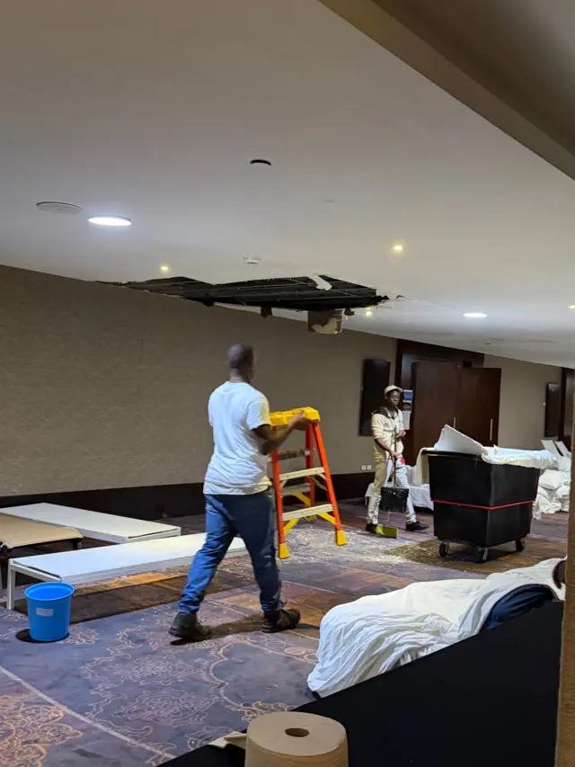 Two hotel workers dressed in white collect bits of ceilings in a laundry bin