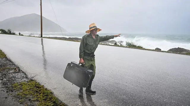 A man holds his hand out and tries to a hitch a ride while holding a suitcase