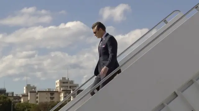Hegseth in a suit and sunglasses walking down the stairs from a plane.