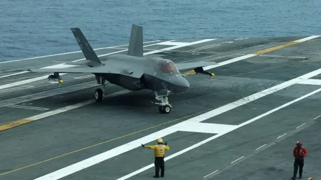 A military jet on the tarmac of an aircraft carrier, with people walking near it