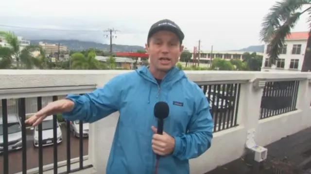 Jason Allen stands on a balcony overlooking a windy, grey landscape with some cars, trees, and power lines in the background. He's wearing a black cap and a blue coat.