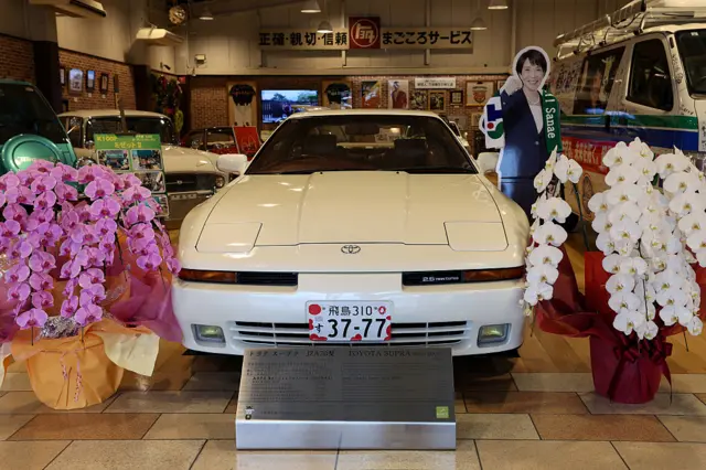 A white Toyota sports car on display beside a cardboard poster of Japanese PM Sanae Takaichi
