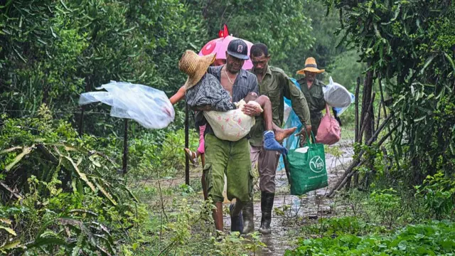 Cubans carry their supplies through a dense forest under rain. One man carries an older person.