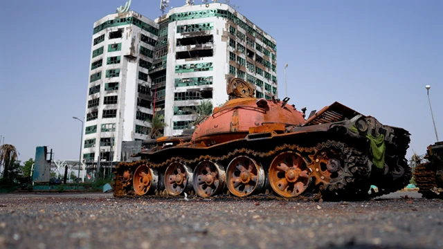 A wrecked tank sits in front of a ruined building